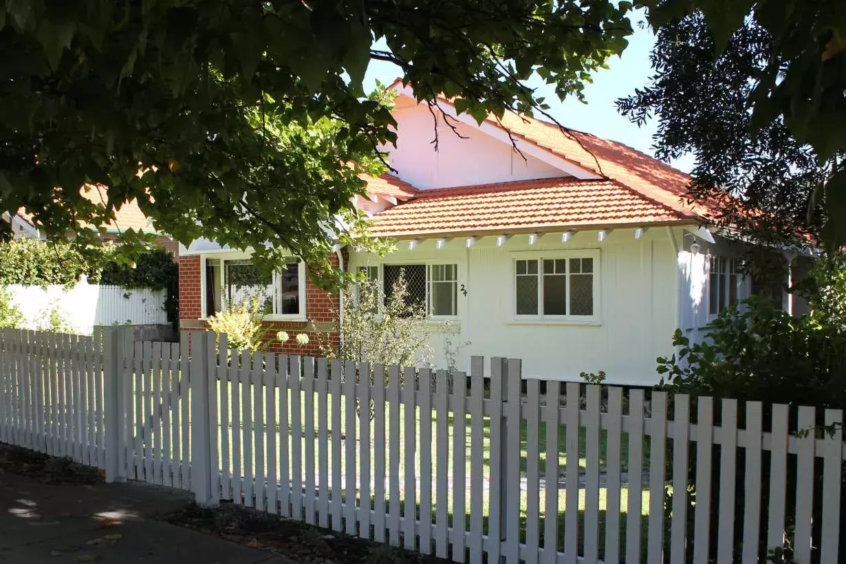 A small white house with a red tile roof with large trees overhanging and white picket fence.  