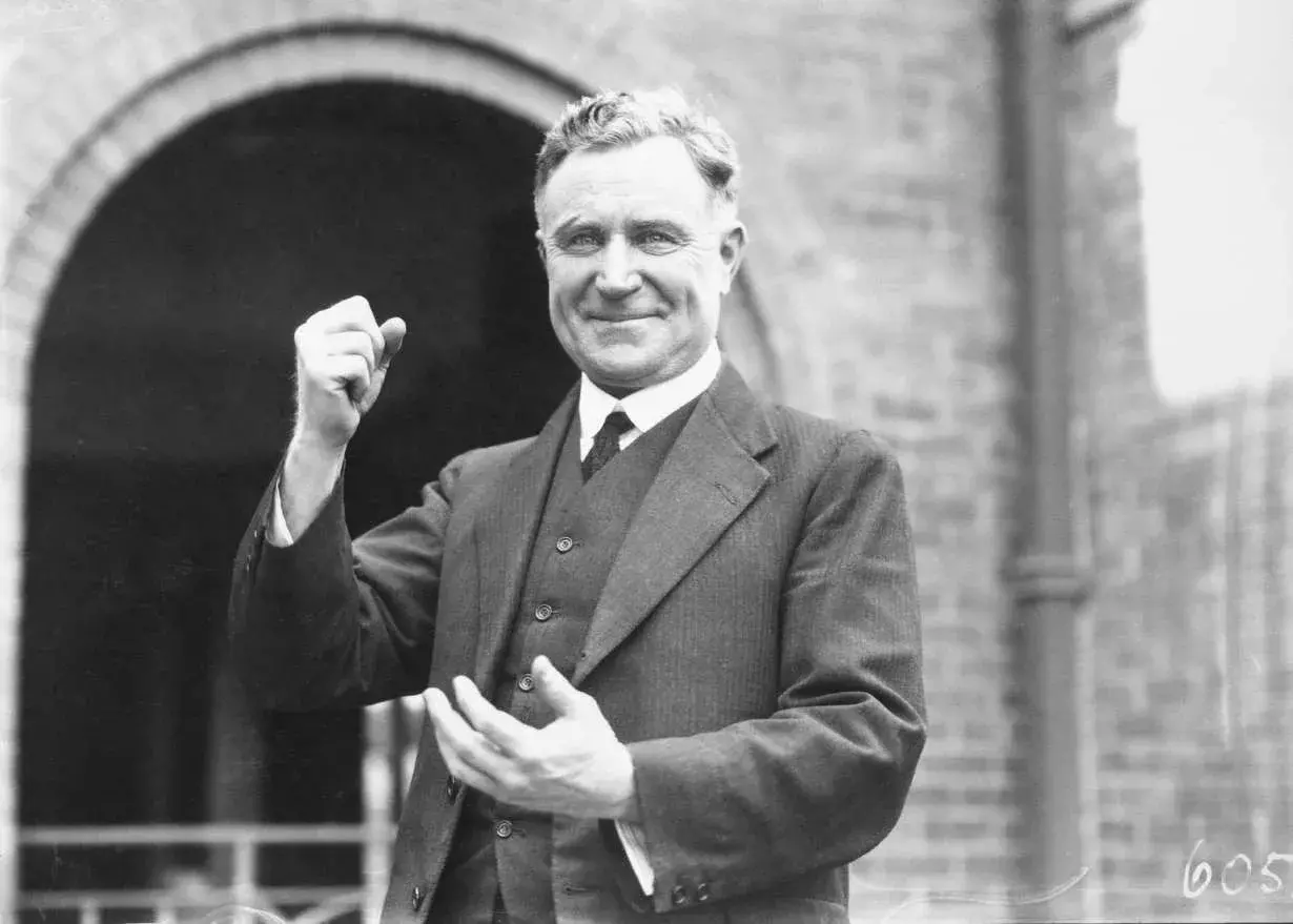 Earle Page standing in front of an archway made of brick, holding his hand up in a gentle fist.  